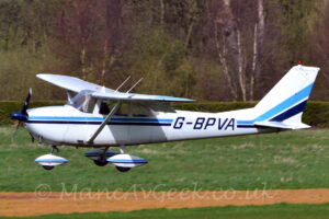 Side view of a high-winged, single propellor-engined light aircraft flying straight and level from right to left at a very low altitude, with flaps extended from the rear of the wings, suggesting it is about to land. The plane is white, with a blue and black stripe running along the body, overlaid with the black registration "G-BPVA" on the sides of the rear fuselage, with similar stripes running over the spats on the undercarriage. Green grass fills the bottom of the frame, except for a narrow strip of yellow grass that runs across the frame. Trees fill the background.