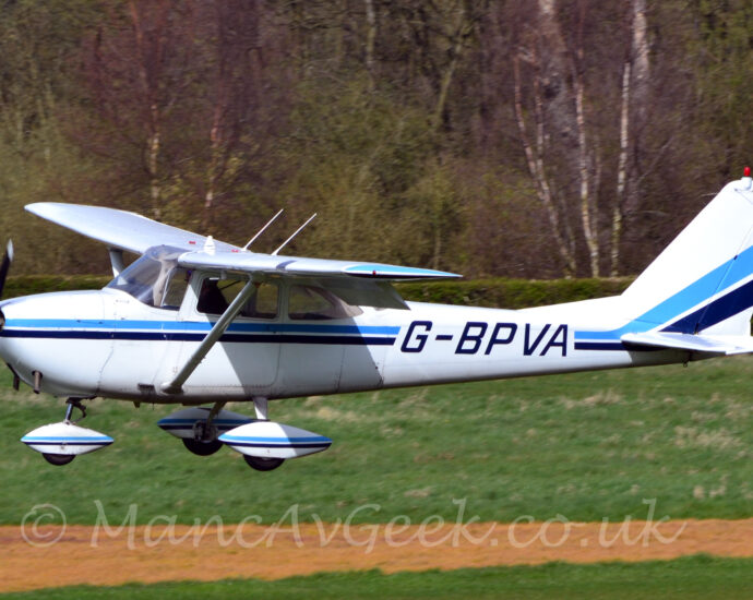 Side view of a high-winged, single propellor-engined light aircraft flying straight and level from right to left at a very low altitude, with flaps extended from the rear of the wings, suggesting it is about to land. The plane is white, with a blue and black stripe running along the body, overlaid with the black registration "G-BPVA" on the sides of the rear fuselage, with similar stripes running over the spats on the undercarriage. Green grass fills the bottom of the frame, except for a narrow strip of yellow grass that runs across the frame. Trees fill the background.