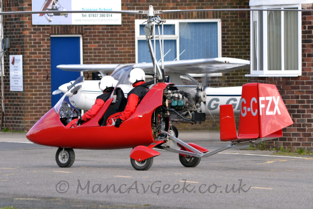 Rear side view of a red, 2 seat autogyro taxiing from right to left with the main rotor locked in place. It is basically a red fibreglass shell wrapped around a metal frame, with a pair of open tandem cockpits with large perspex windshields, with a motor bolted on the back powering a pusher propellor, and another metal frame sticking backwards out of the bottom carrying the red tail on an angular metal boom. An uncovered nosewheel sticks out of the bottom of the fuselage, with a pair of rear wheel on struts, partially covered with red sponsons stick out to the sides from under the engine. The silver registration is on the red tail right at the back.Several metal poles stick out from the engine, just above the rear cockpit, supporting and providing some control to the large, 2-bladed main rotor. Both cockpit seats are filled, with people wearing matching flying suits, with red arms and legs and black bodies, as well as white full-face helmets. Grey tarmac taxiway fills the foreground, with 2 wings of an old, brown brick office building in the background, close enough to possibly be hit if the main rotor was spinning, with a white, high-winged, light aircraft parked between the wings.
