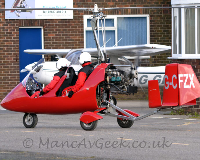Rear side view of a red, 2 seat autogyro taxiing from right to left with the main rotor locked in place. It is basically a red fibreglass shell wrapped around a metal frame, with a pair of open tandem cockpits with large perspex windshields, with a motor bolted on the back powering a pusher propellor, and another metal frame sticking backwards out of the bottom carrying the red tail on an angular metal boom. An uncovered nosewheel sticks out of the bottom of the fuselage, with a pair of rear wheel on struts, partially covered with red sponsons stick out to the sides from under the engine. The silver registration is on the red tail right at the back.Several metal poles stick out from the engine, just above the rear cockpit, supporting and providing some control to the large, 2-bladed main rotor. Both cockpit seats are filled, with people wearing matching flying suits, with red arms and legs and black bodies, as well as white full-face helmets. Grey tarmac taxiway fills the foreground, with 2 wings of an old, brown brick office building in the background, close enough to possibly be hit if the main rotor was spinning, with a white, high-winged, light aircraft parked between the wings.