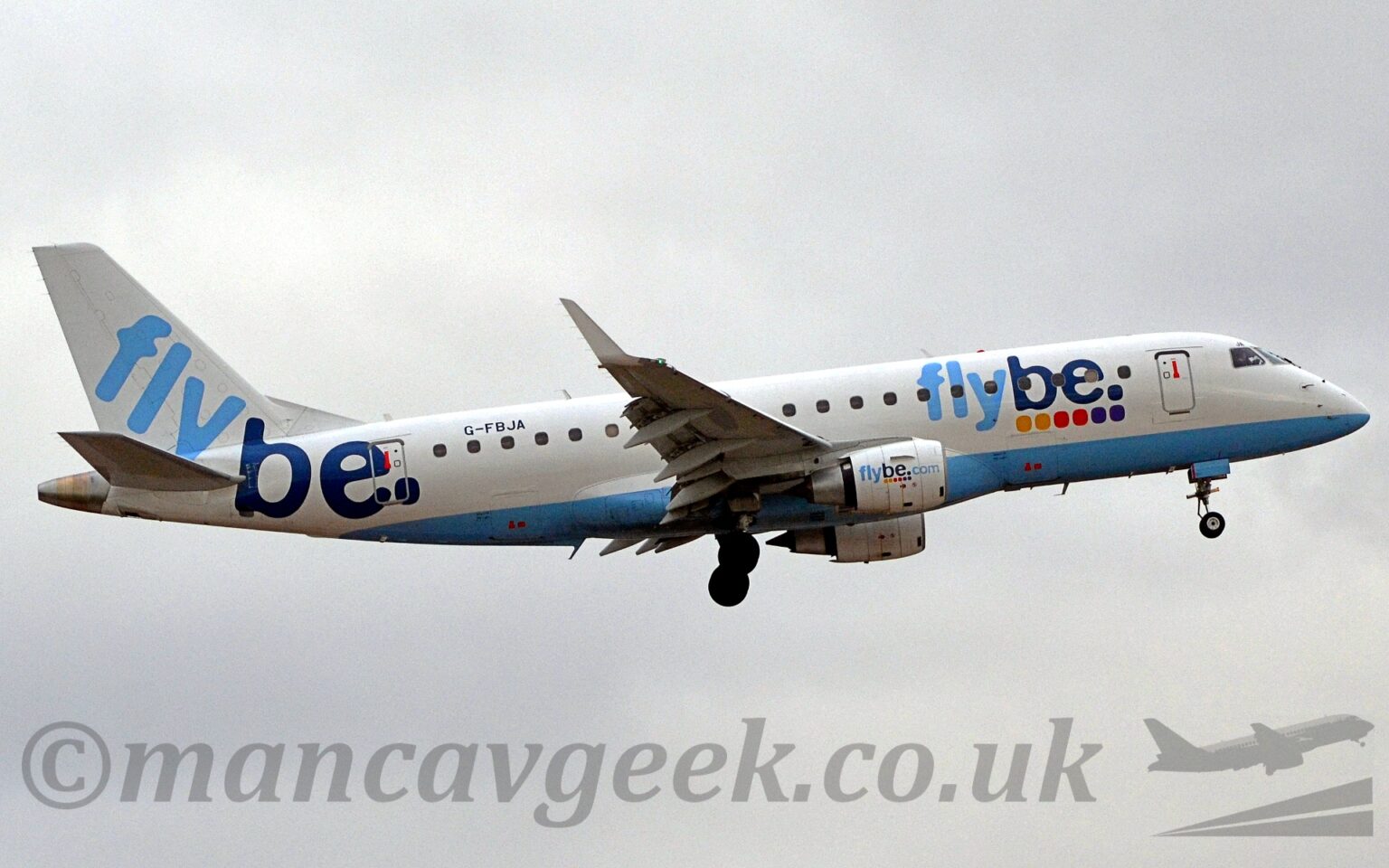 Side view of a twin engined jet airliner flying from left to right at a very low altitude, with undercarriage lowered and flaps deployed from the rear of the wings, suggesting it is about to land. The plane is mostly white, with a pale blue belly, and two-tone light and dark blue "FlyBe" titles on the side of the forward fuselage, over pairs of yellow, red, and purple dots. The same logo appears in a smaller form on the sides of the white engine pods under the wings. A larger version of the logo, without the dots, appears diagonally on the white tail and rear fuselage, just aft of the black registration "G-FBJA". Flat grey sky fills the rest of the frame.