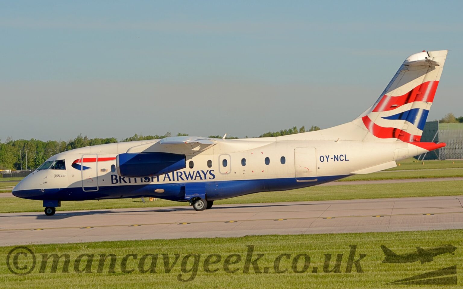 Side view of a high-winged, twin engined jet airliner lined up waiting for take off on a grey runway. The plane is largely white with a dark blue belly, and dark blue "British Airways" titles on the lower forward fuselage. A twisted, red and blue ribbon is on the upper forward fuselage, immediately aft of the cockpit windows, and the black registration "OY-NCL" is on the upper rear fuselage. The tail is white, with rippling red stripes either side of a rippling dark blue triangle in the middle. The horizontal stabiliser, mounted at the top of the tail, is casting it's own shadow on the top and front of the tail, which suggests that the sun is up in the top left corner somewhere. Green grass fills the foreground, lining both sides of the runway. Trees in the distance meet grey clouds and blue sky.