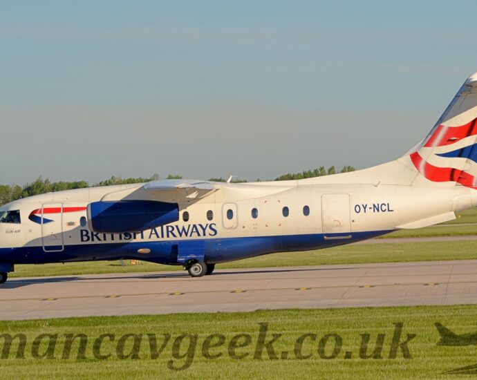 Side view of a high-winged, twin engined jet airliner lined up waiting for take off on a grey runway. The plane is largely white with a dark blue belly, and dark blue "British Airways" titles on the lower forward fuselage. A twisted, red and blue ribbon is on the upper forward fuselage, immediately aft of the cockpit windows, and the black registration "OY-NCL" is on the upper rear fuselage. The tail is white, with rippling red stripes either side of a rippling dark blue triangle in the middle. The horizontal stabiliser, mounted at the top of the tail, is casting it's own shadow on the top and front of the tail, which suggests that the sun is up in the top left corner somewhere. Green grass fills the foreground, lining both sides of the runway. Trees in the distance meet grey clouds and blue sky.