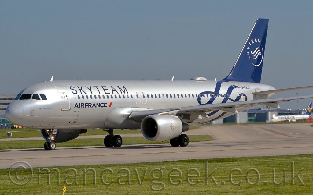 Side view of a twin engined jet airliner taxiing from right to left, turning on to a grey runway. The plane is in a silver finish, with dark blue "SkyTeam" titles on the upper forward fuselage, above the forward cabin windows, and smaller "Air France" titles under those windows. The rear fuselage has some dark blue scrollwork, next to the black registration "F-GKXS" on the upper rear fuselage. The dark blue tail has a thin crescent forming the bottom of a white circle, the white word "SkyTeam" forming the top, and has some more white scrollwork in the middle. Green grass lines the sides of the taxiway in the foreground, with blue-grey sky filling the rest of the frame.