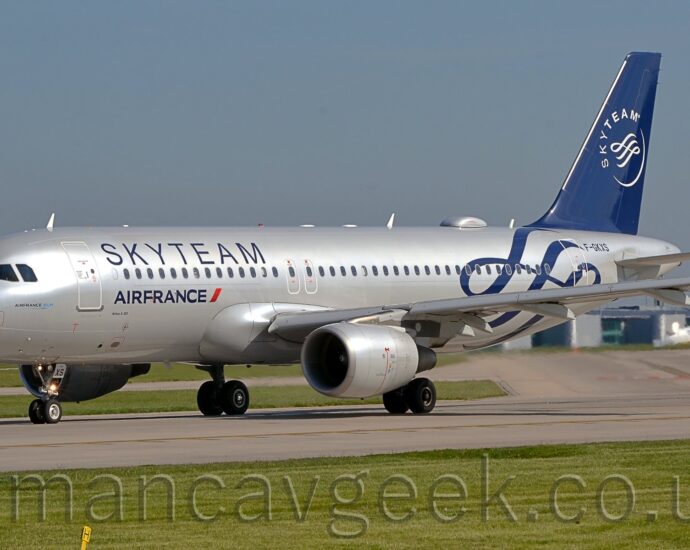 Side view of a twin engined jet airliner taxiing from right to left, turning on to a grey runway. The plane is in a silver finish, with dark blue "SkyTeam" titles on the upper forward fuselage, above the forward cabin windows, and smaller "Air France" titles under those windows. The rear fuselage has some dark blue scrollwork, next to the black registration "F-GKXS" on the upper rear fuselage. The dark blue tail has a thin crescent forming the bottom of a white circle, the white word "SkyTeam" forming the top, and has some more white scrollwork in the middle. Green grass lines the sides of the taxiway in the foreground, with blue-grey sky filling the rest of the frame.