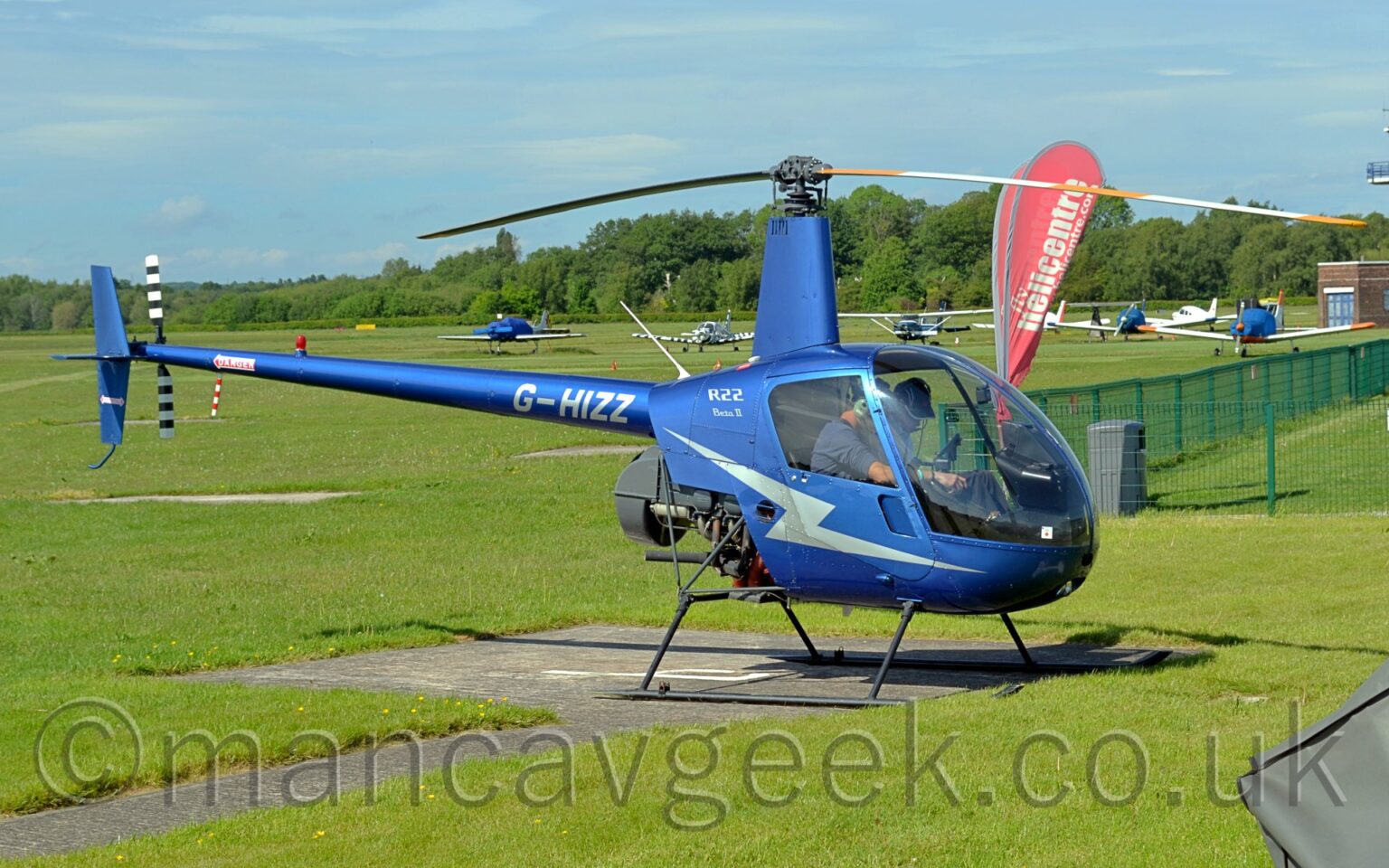 Side view of a metallic blue small helicopter parked on a concrete landing pad, facing to the right. There is a light blue lightning flash on the lower fuselage and door, with silver "R22"text on the upper rear fuselage, above smaller "Beta II" text. The silver registration "G-HIZZ" is at the front of the tail boom, just as it comes out of the fuselage, above the partially exposed engine. The black and white striped tail rotor is on the left side at the rear of the tail boom. Green grass fills the foreground, with several planes parked on more grass in the background, leading up to trees and bushes marking the airfield perimeter in the distance, under blue sky with smatterings of thin white cloud.