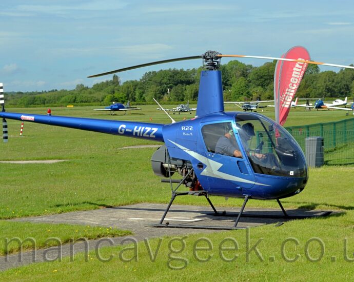 Side view of a metallic blue small helicopter parked on a concrete landing pad, facing to the right. There is a light blue lightning flash on the lower fuselage and door, with silver "R22"text on the upper rear fuselage, above smaller "Beta II" text. The silver registration "G-HIZZ" is at the front of the tail boom, just as it comes out of the fuselage, above the partially exposed engine. The black and white striped tail rotor is on the left side at the rear of the tail boom. Green grass fills the foreground, with several planes parked on more grass in the background, leading up to trees and bushes marking the airfield perimeter in the distance, under blue sky with smatterings of thin white cloud.