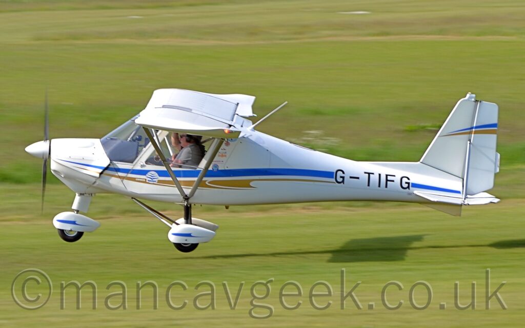 Side-view of a single-engined, high-winged light aircraft flying from right to left at a very low altitude, just centimetres between the grass runway and the plane's wheels. The plane is largely white, with a blue and brown stripe running along the body, overlaid with the black registration "G-TIFG" on the side of the rear fuselage. White spats cover all 3 wheels, with blue stripe running along each one2 people can be seen i the rather cramped, fully glazed cockpit. The plane is casting a strong shadow on the ground quite a way behind it, suggesting that the sun is about 3 quarters of the way up the left of the frame. Green grass fills the rest of the image.