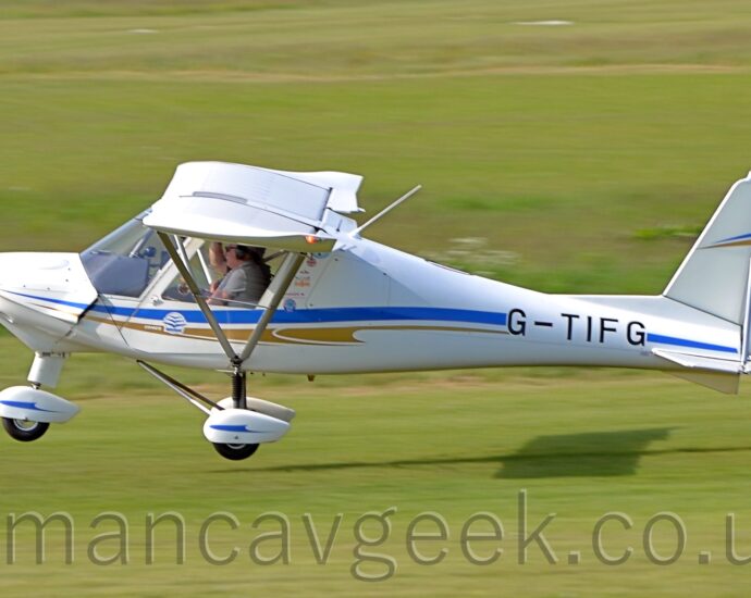 Side-view of a single-engined, high-winged light aircraft flying from right to left at a very low altitude, just centimetres between the grass runway and the plane's wheels. The plane is largely white, with a blue and brown stripe running along the body, overlaid with the black registration "G-TIFG" on the side of the rear fuselage. White spats cover all 3 wheels, with blue stripe running along each one2 people can be seen i the rather cramped, fully glazed cockpit. The plane is casting a strong shadow on the ground quite a way behind it, suggesting that the sun is about 3 quarters of the way up the left of the frame. Green grass fills the rest of the image.