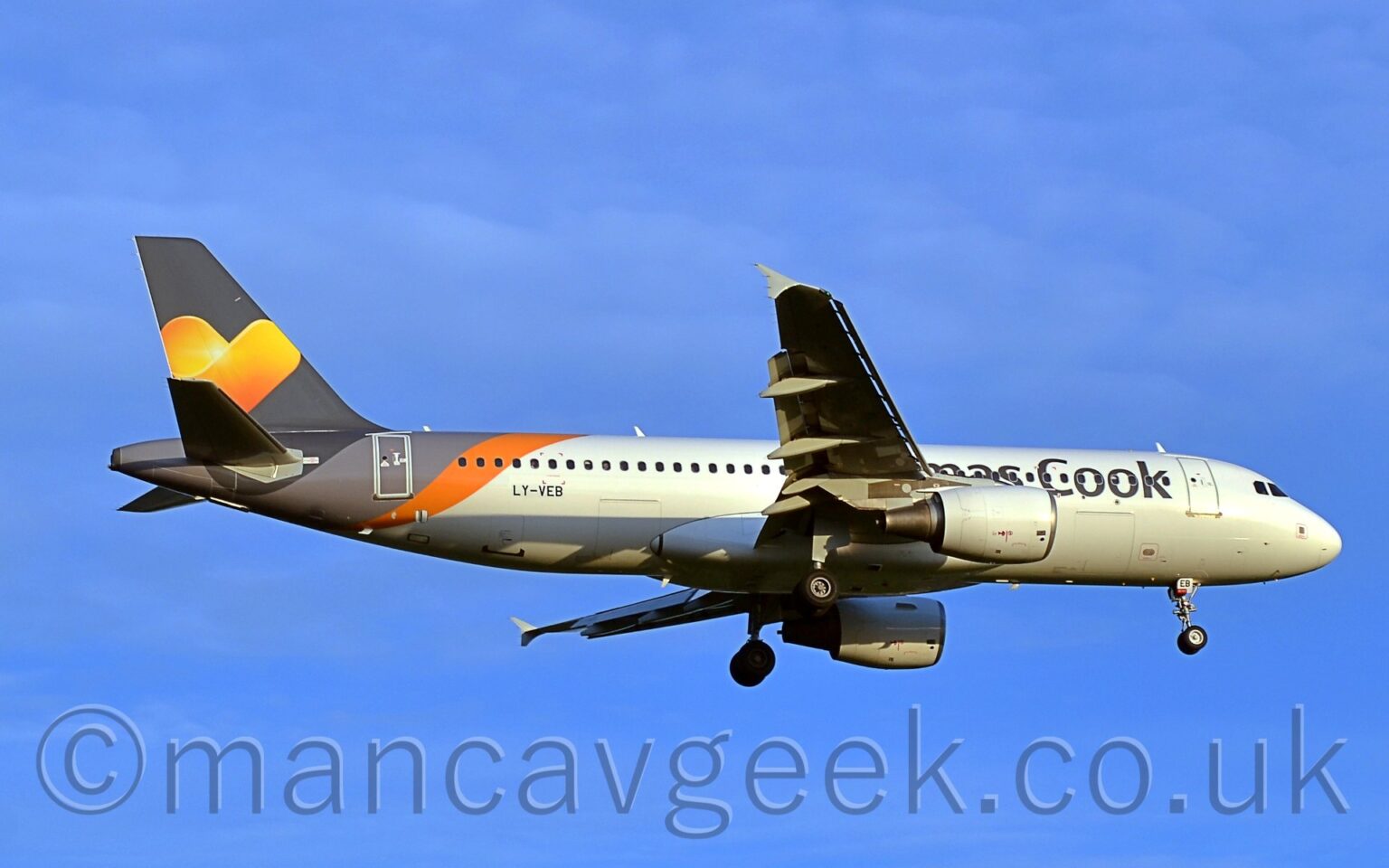 Side view of a twin engined jet airliner flying from left to right at a low altitude, with undercarriage lowered and flaps extended from the rear of the wings, suggesting it is about to land. The planes id largely white with partially obscured "Thomas Cook" titles on the side of the forward fuselage, and the black registration "LY-VEB" on the lower rear fuselage. A diagonal light grey and orange band covers the rear fuselage, below the grey tail, which has a large yellow and orange love heart in the middle. Vivid blue sky with occasional light fluffs of thin cloud fills the background.