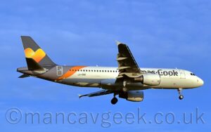Side view of a twin engined jet airliner flying from left to right at a low altitude, with undercarriage lowered and flaps extended from the rear of the wings, suggesting it is about to land. The planes id largely white with partially obscured "Thomas Cook" titles on the side of the forward fuselage, and the black registration "LY-VEB" on the lower rear fuselage. A diagonal light grey and orange band covers the rear fuselage, below the grey tail, which has a large yellow and orange love heart in the middle. Vivid blue sky with occasional light fluffs of thin cloud fills the background.