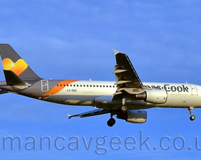 Side view of a twin engined jet airliner flying from left to right at a low altitude, with undercarriage lowered and flaps extended from the rear of the wings, suggesting it is about to land. The planes id largely white with partially obscured "Thomas Cook" titles on the side of the forward fuselage, and the black registration "LY-VEB" on the lower rear fuselage. A diagonal light grey and orange band covers the rear fuselage, below the grey tail, which has a large yellow and orange love heart in the middle. Vivid blue sky with occasional light fluffs of thin cloud fills the background.