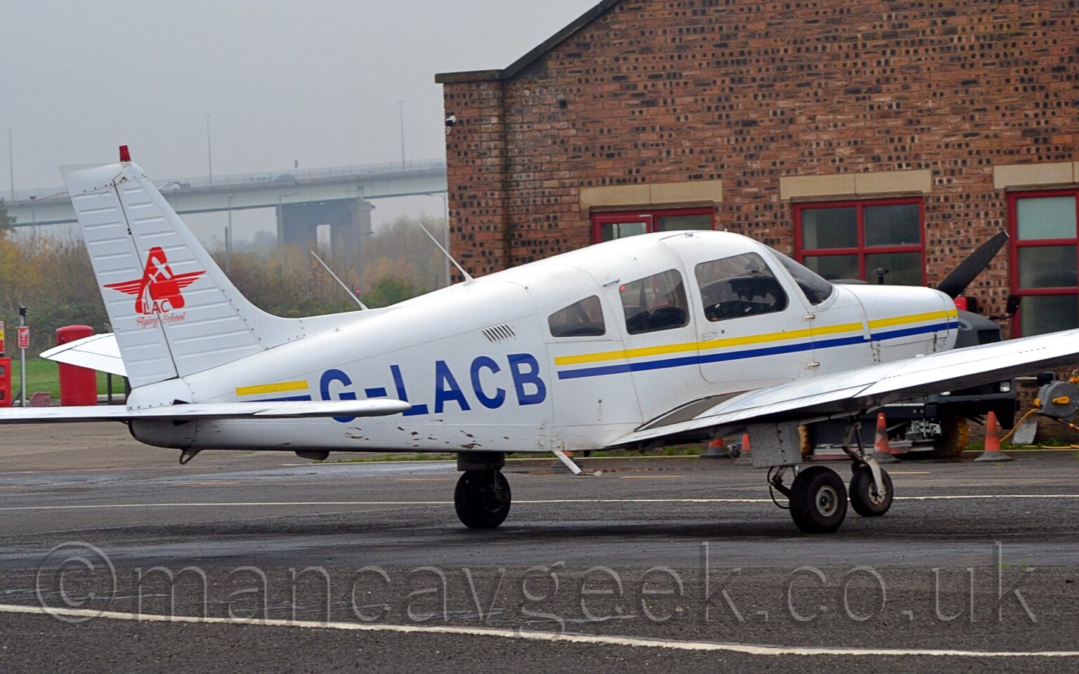 Rear side view of a single engined light aircraft parked facing to the right on black tarmac. The plane is largely white, with a thin blue and yellow stripe running along the body, overlaid with the black registration "G-LACB" on the side of the rear fuselage. There is a red star on the white tail, made up of an upper case letter "A" with wings at the sides, overlaid with the silhouette of a small plane flying upwards, and with the text "LAC Flying School" at the bottom. Black tarmac fills the foreground, with a red brick building in the background, on the right, and trees on the left, with a tall bridge in the distance, under flat grey sky.