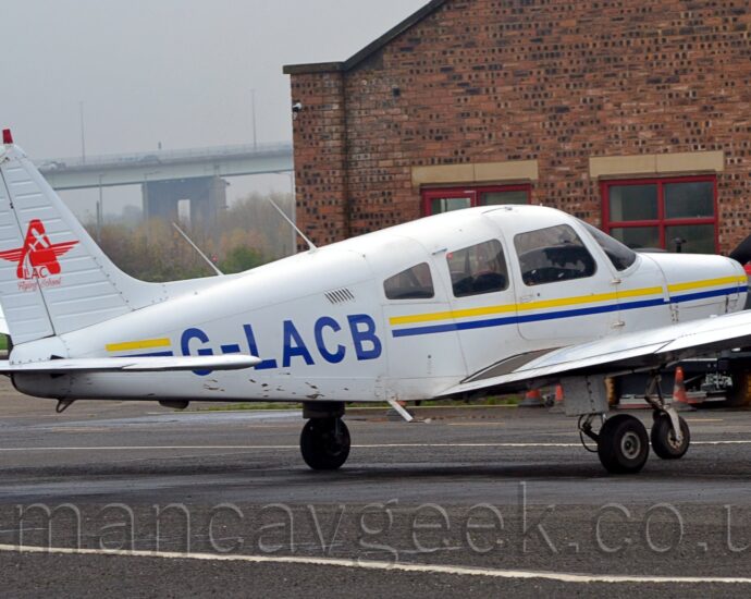 Rear side view of a single engined light aircraft parked facing to the right on black tarmac. The plane is largely white, with a thin blue and yellow stripe running along the body, overlaid with the black registration "G-LACB" on the side of the rear fuselage. There is a red star on the white tail, made up of an upper case letter "A" with wings at the sides, overlaid with the silhouette of a small plane flying upwards, and with the text "LAC Flying School" at the bottom. Black tarmac fills the foreground, with a red brick building in the background, on the right, and trees on the left, with a tall bridge in the distance, under flat grey sky.