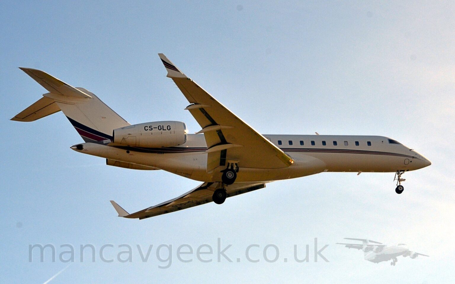 Low side view of a white, twin engined Bizjet flying from left to right at a very low altitude. The plane has it's undercarriage lowered, and flaps extended from the rear of the wings, suggesting it is about to land. The plane is mostly white, with a dark blue, red, and grey stripe running along the body from the nose, getting slightly wider and sweeping up in to the lower reaches of the tail. A similar stripe is on the outer face of the up-turned wing-tips. Bright but hazy sky with hints of blue fill the rest of the frame.