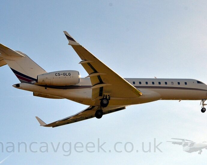 Low side view of a white, twin engined Bizjet flying from left to right at a very low altitude. The plane has it's undercarriage lowered, and flaps extended from the rear of the wings, suggesting it is about to land. The plane is mostly white, with a dark blue, red, and grey stripe running along the body from the nose, getting slightly wider and sweeping up in to the lower reaches of the tail. A similar stripe is on the outer face of the up-turned wing-tips. Bright but hazy sky with hints of blue fill the rest of the frame.