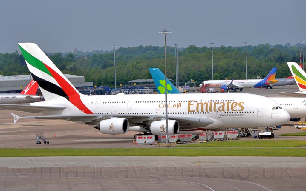 Side view of a very large, 4 engined jet airliner parked facing to the right on a large section of concrete apron. The plane is almost entirely white, with large, golden "Emirates" titles on the forward fuselage, between the 2 passenger deck windows. A large, diagonal version of the United Arab Emirates flag fills the tail and part of the upper rear fuselage. Grey taxiways and a strip of green grass fills the foreground, with several more jet airliners parked in the background, with trees in the distance, under grey sky.