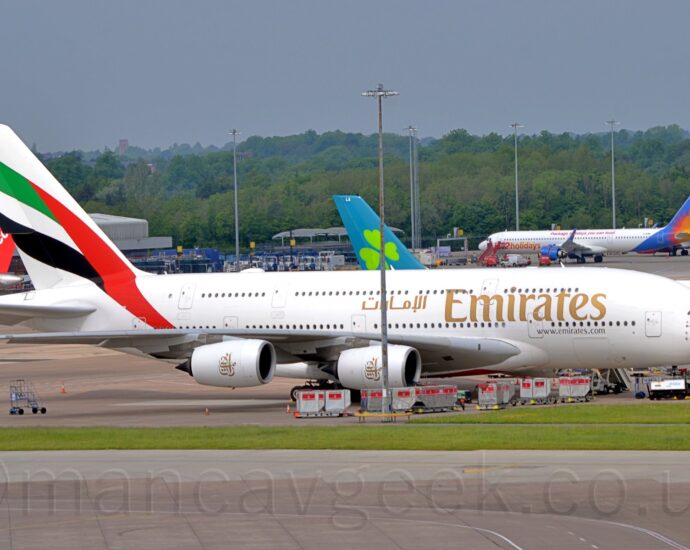 Side view of a very large, 4 engined jet airliner parked facing to the right on a large section of concrete apron. The plane is almost entirely white, with large, golden "Emirates" titles on the forward fuselage, between the 2 passenger deck windows. A large, diagonal version of the United Arab Emirates flag fills the tail and part of the upper rear fuselage. Grey taxiways and a strip of green grass fills the foreground, with several more jet airliners parked in the background, with trees in the distance, under grey sky.