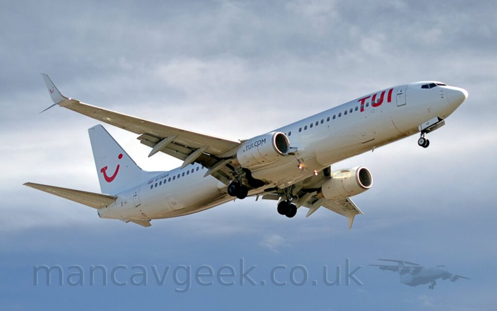 Side view of a white, twin engined jet airliner flying from left to right, with undercarriage lowered and flaps extended from the rear of the wings, suggesting it is about to land.. The plane has red "TUI" titles on the upper forward fuselage, and a red logo on that tail that appears to be a mix of the letters "T", "U", and "I", and a winking smiley face. There are blue "TUI[dot]com" titles on the sides of the white engine pods. The registration "G-TUKR" is visible on the upper rear fuselage, next to a small British flag. Cloudy grey sky with patches of blue fills the rest of the frame.