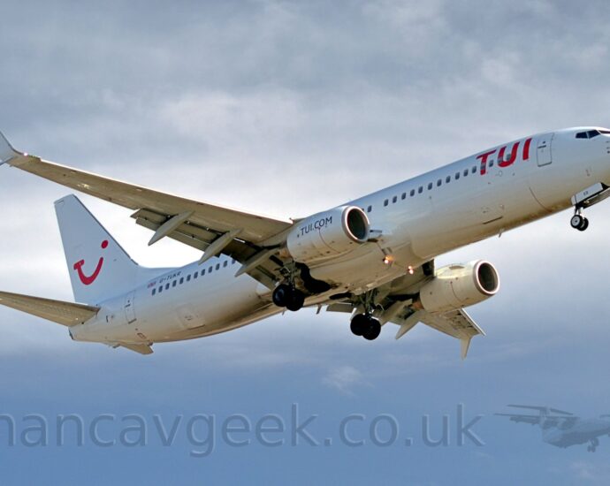 Side view of a white, twin engined jet airliner flying from left to right, with undercarriage lowered and flaps extended from the rear of the wings, suggesting it is about to land.. The plane has red "TUI" titles on the upper forward fuselage, and a red logo on that tail that appears to be a mix of the letters "T", "U", and "I", and a winking smiley face. There are blue "TUI[dot]com" titles on the sides of the white engine pods. The registration "G-TUKR" is visible on the upper rear fuselage, next to a small British flag. Cloudy grey sky with patches of blue fills the rest of the frame.