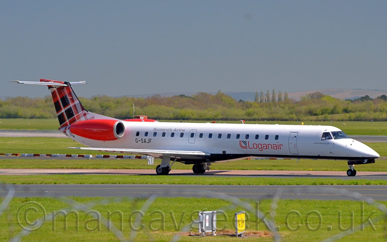 Side view of a twin engined jet airliner with the engines mounted on the sides of the rear fuselage, taxiing from left to right along a reddish-grey taxiway. The plane is mostly white, with red "LoganAir" titles on the lower forward fuselage, and the black registration "G-SAJF" on the lower rear, just below and forward of the red engine pods. The tail is covered in a black, red, and white tartan pattern. Green grass fills the foreground, with some out-of-focus rolls of razor wire at the bottom of the frame. More grass in the background leads up to trees in the distance, under grey-blue skies.
