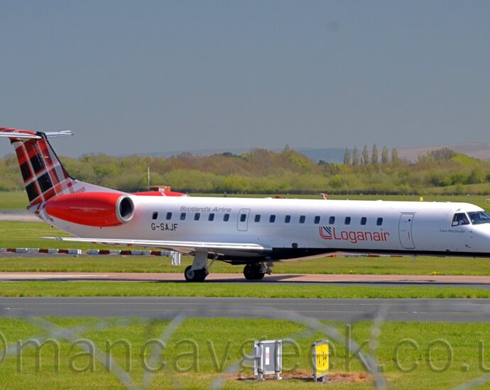 Side view of a twin engined jet airliner with the engines mounted on the sides of the rear fuselage, taxiing from left to right along a reddish-grey taxiway. The plane is mostly white, with red "LoganAir" titles on the lower forward fuselage, and the black registration "G-SAJF" on the lower rear, just below and forward of the red engine pods. The tail is covered in a black, red, and white tartan pattern. Green grass fills the foreground, with some out-of-focus rolls of razor wire at the bottom of the frame. More grass in the background leads up to trees in the distance, under grey-blue skies.