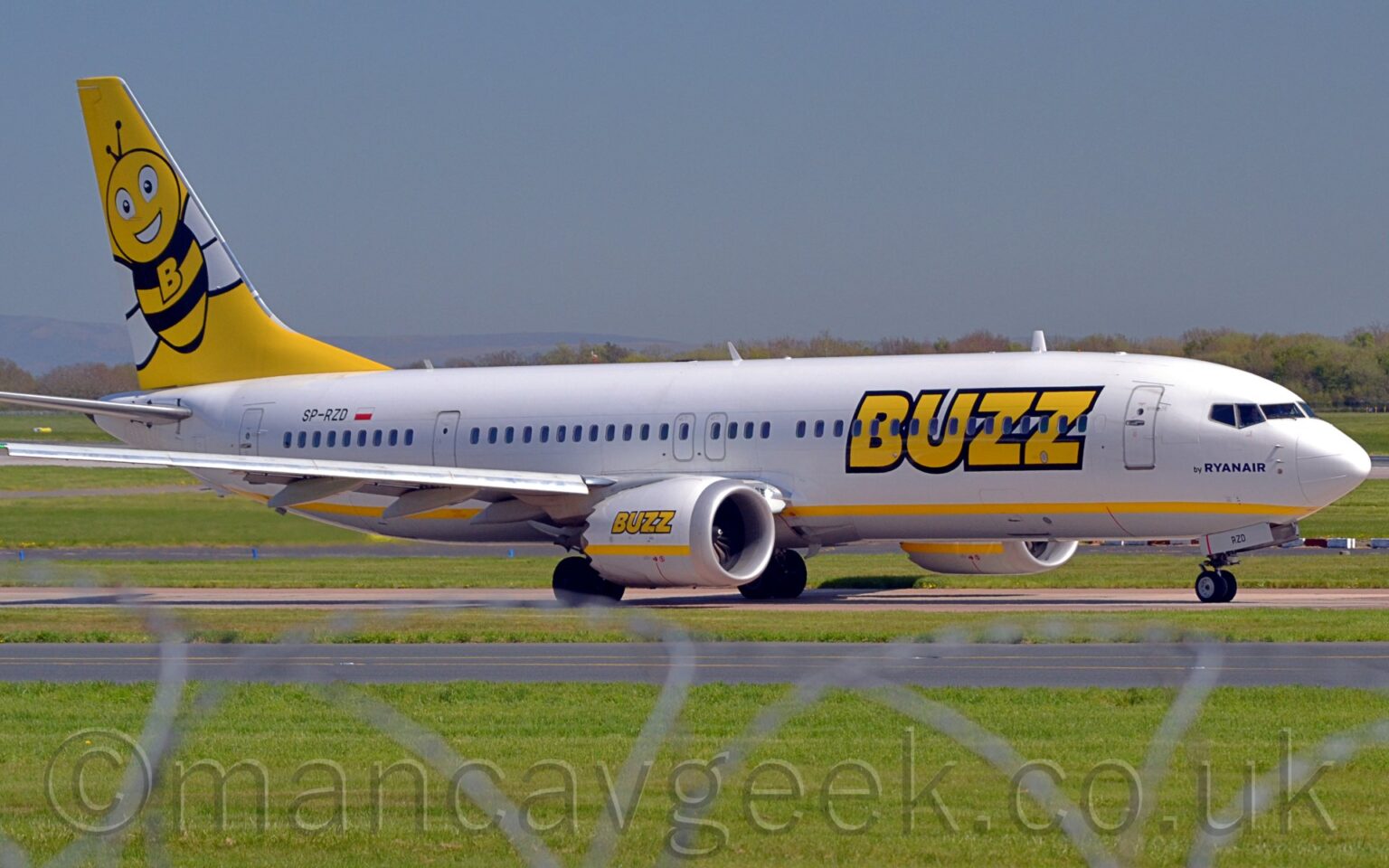 Side view of a twin engined jet airliner taxiing from left to right along a grey taxiway. The plane is mostly white, with a yellow stripe running along the lower body, and large yellow "Buzz" titles outlined in black on the forward fuselage. These titles are repeated over a yellow stripe on the sides of the engine pods, under the wings. There is dark blue text "by RyanAir" under the cockpit windows, and the black registration "SP-RZD" just aft of a white and red Polish flag on the upper rear fuselage. The tail is bright yellow, with a large cartoon image of a bee, with a large upper-case letter "B" on it's black and yellow striped chest. Green grass fills the foreground, partially obscured by a stretched-out roll of razorwire on top. More grass in the background leads up to trees in the distance, under blue sky.