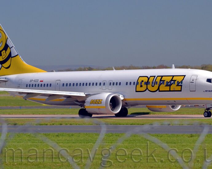 Side view of a twin engined jet airliner taxiing from left to right along a grey taxiway. The plane is mostly white, with a yellow stripe running along the lower body, and large yellow "Buzz" titles outlined in black on the forward fuselage. These titles are repeated over a yellow stripe on the sides of the engine pods, under the wings. There is dark blue text "by RyanAir" under the cockpit windows, and the black registration "SP-RZD" just aft of a white and red Polish flag on the upper rear fuselage. The tail is bright yellow, with a large cartoon image of a bee, with a large upper-case letter "B" on it's black and yellow striped chest. Green grass fills the foreground, partially obscured by a stretched-out roll of razorwire on top. More grass in the background leads up to trees in the distance, under blue sky.