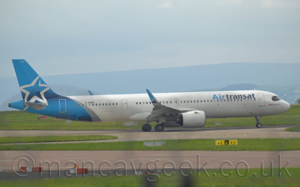Side view of a twin engined jet airliner taxiing from left to right along a grey taxiway. The plane is largely white, with light and dark blue "Air Transat" titles on the upper forward fuselage, with the black registration "C-GOIM" on the upper rear. The tail and rear fuselage are light blue, with a diagonal silver band separating the blue and the white. The outline of a large silver star adorns the tail. Large grassed areas and grey taxiways fill the foreground, with rolling grey hills in the background, under blue-grey sky.
