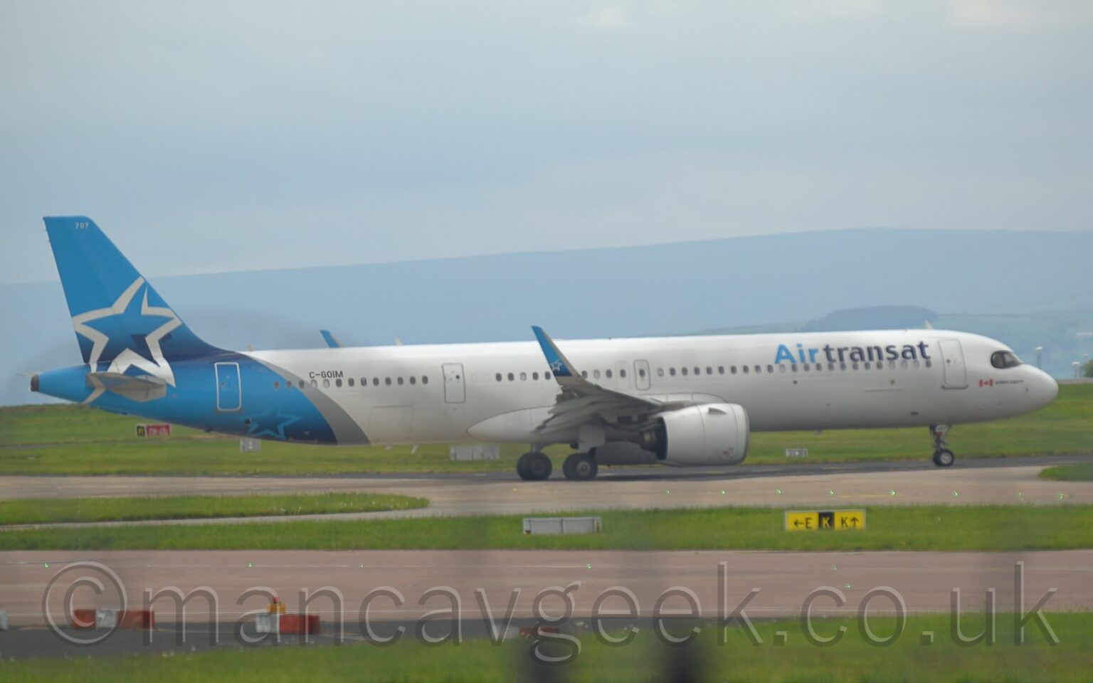 Side view of a twin engined jet airliner taxiing from left to right along a grey taxiway. The plane is largely white, with light and dark blue "Air Transat" titles on the upper forward fuselage, with the black registration "C-GOIM" on the upper rear. The tail and rear fuselage are light blue, with a diagonal silver band separating the blue and the white. The outline of a large silver star adorns the tail. Large grassed areas and grey taxiways fill the foreground, with rolling grey hills in the background, under blue-grey sky.