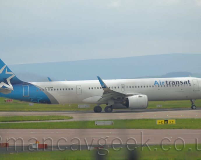 Side view of a twin engined jet airliner taxiing from left to right along a grey taxiway. The plane is largely white, with light and dark blue "Air Transat" titles on the upper forward fuselage, with the black registration "C-GOIM" on the upper rear. The tail and rear fuselage are light blue, with a diagonal silver band separating the blue and the white. The outline of a large silver star adorns the tail. Large grassed areas and grey taxiways fill the foreground, with rolling grey hills in the background, under blue-grey sky.