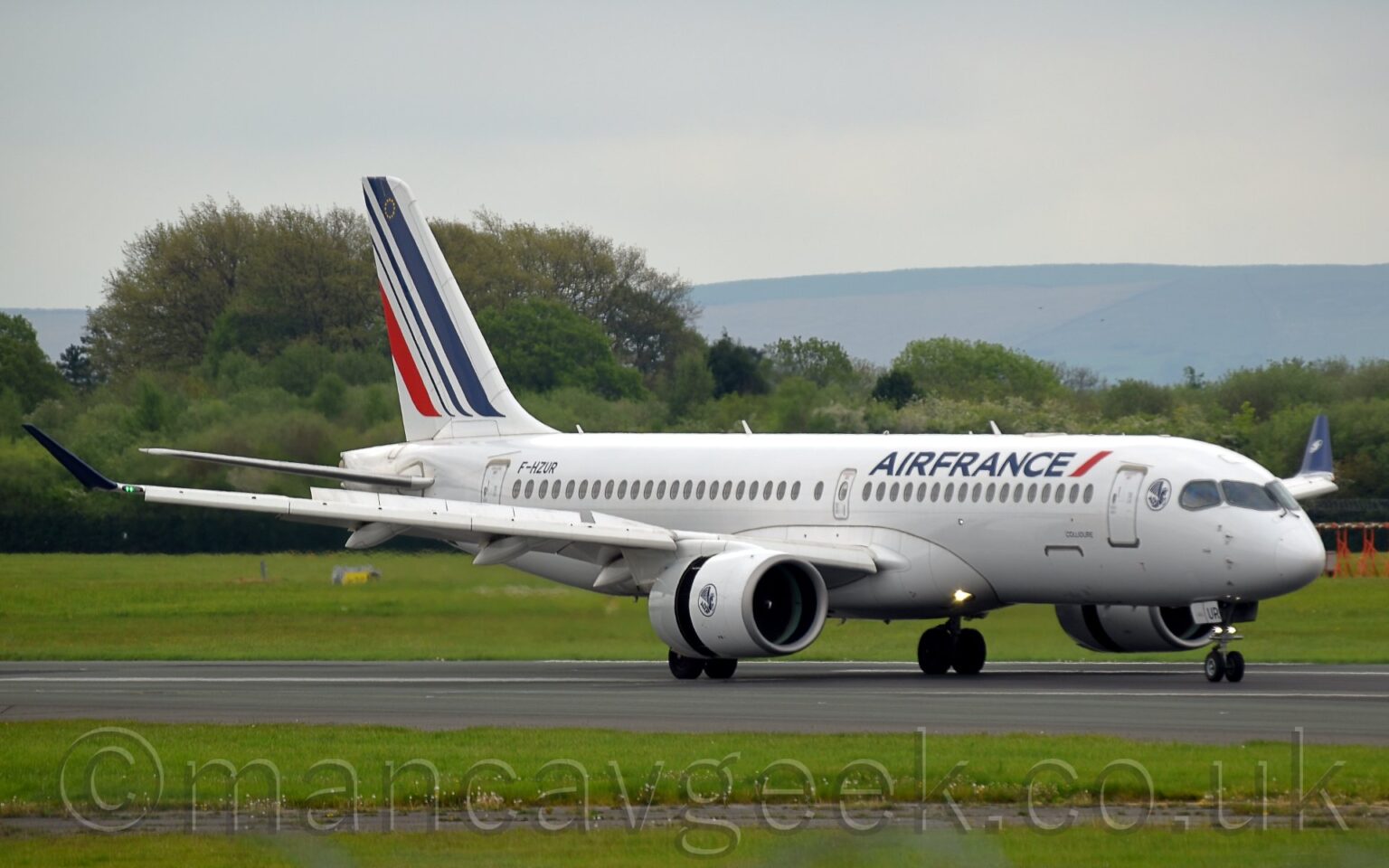 Front side view of a white, twin engined jet airliner moving from left to right along a black runway, with flaps extended from the rear of the wings, and the rear section of the white engine pods pushed backwards, revealing the thrust reversers, suggesting it has just landed. There are dark blue "Air France" titles on the upper forward fuselage, next to a short, red diagonal stripe, and the black registration "F-HZUR" on the upper rear. There is an image of a dark blue winged sea horse in the outline of a blue circle on the side of the front section of the white engine pods under the wings. 3 diagonal blue stripes, getting thinner as they move rearwards, followed by a shorter red stripe, adorn the tail, with a small circle of gold stars at the top. Green grass lines the side of the runway in the foreground, with more grass in the background leading up to trees in the distance, with rolling hills beyond that, under pale blue sky.