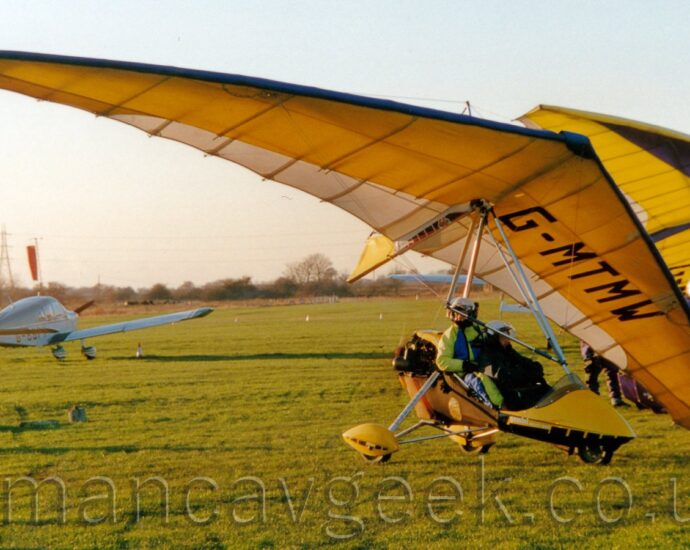 Front side view of a microlight aircraft parked on grass, facing to the right, with its left wingtip resting on the ground. What little fuselage there is, is a metal frame covered in a carbon fibre shell, with a pair of tandem seats in an open cockpit on top, with a small engine driving a pusher propellor immediately behind, suspended under a large, triangular parasol wing. A small nosewheel is embedded in the forward shell, with main gear covered in yellow spats are on the end of outriggers attached just in front of the engine. The carbon fibre shell is bright yellow, with the wing being mostly yellow, and blue leading edges and cream trailing edge. The black registration "G-MTMW" is on the underside of the left side of the wing. Green grass fills most of the bottom of the frame, with another microlight with a yellow and purple wing on the right of the background, and a single engined light aircraft taxiing away from the camera on the left. Bright but hazy sky fills the rest of the frame.