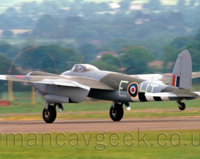Side view of a twin propellor-engined, World War 2-era fighter-bomber accelerating from right to left along a grey tarmac runway, with it's tail wheel raised off the ground, suggesting it is taking off. The plane has a grey and olive green camouflage scheme on the upper body and upper wing surfaces, with grey belly and undersides of the wings. There are 3 white and 2 black bands under the lower rear fuselage. The pale mint green letters "E-HT" are either side of a blue, white, and red roundel, outlined in yellow, on the rear fuselage, with the black serial "RR299" just after, under a red, white, and blue fin-flash on the tail. Green grass fills the foreground, with trees on small hills in the background.