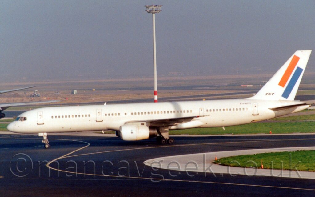 Side view of a white, twin engined jet airliner in a lightly modified "Polar bear in a snow storm" livery - almost entirely white, with only the black registration "PH-AHE" on the upper rear fuselage, black text "Boeing 757"at the base of the tail, and diagonal orange and blue stripes on the tail itself marring the bland uniformity. A black taxiway with a yellow centreline winds its way through the frame, around concrete islands with grassy interiors in the foreground and through the background. A tall lighting pole reaches into the sky in the middle of the image. Grey-blue sky fills the rest of the frame.