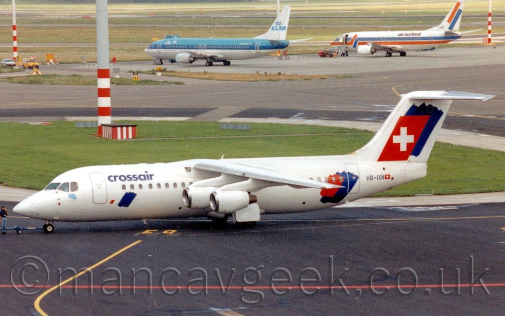 Side view of a white, high-winged, 4 engined jet airliner facing to the left but being pushed back to the right from it's stand . The plane is mostly white, with blue "CrossAir" titles on the upper forward fuselage, and a diagonal blue splodge below the forward cabin widows. A similar but larger blue splodge is on the rear fuselage, partially overlaid with a slightly smaller red splodge, and again, larger still on the white tail, where it is overlaid with a red quadrilateral with a large white cross in the middle, representing the Swiss flag. The black registration "HB-IXN" is on the rear fuselage, next to a small Swiss flag. A long blue pole is connected to the nosewheel on the left of the frame, the other end attached to a tug that is out of frame. Black apron marked with red and yellow lines fills the foreground, with black and grey taxiway stretching across the frame, with a pair of jet airliner, one blue and white, the other white with an orange and blue stripe, with grass beyond them. A few tall lighting poles seem to grow from islands of grass, red and white stripes at the bottom, stretching up and out of the frame.