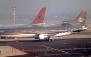 Side view of a 3 engined jet airliner taxiing from right to left along a black taxiway. The plane is a dark purple, almost black, on it's upper half, and a creamy grey on the lower section, with a thin red and gold stripe running along the border between the two colours. There are gold "Royal Jordanian" titles on the upper forward fuselage, in English towards the front, and in an ornate Arabic script above the wing root. There is a large golden crown on the dark purple tail, which also has a thick red stripe right at the top. Black tarmac apron fills the foreground, with the white fuselage and orange tail of a very large, 4 engined jet airliner in the background on the left, and trees vanishing into hazy sky in the far distance.