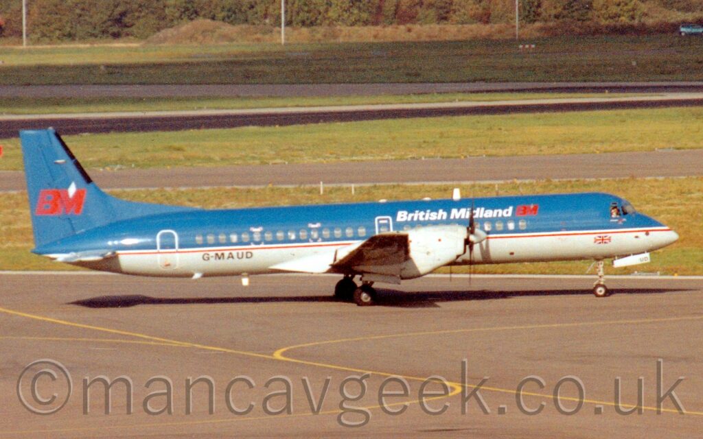 Side view of a twin propellor-engined airliner taxiing from left to right along a grey taxiway. Yellow lines mark the centre line, but this plane isn't following any of them. The plane is dark blue on top and pale grey on the bottom, with white "British Midland" titles on the upper forward fuselage, and a red "BM" just ahead of that, repeated in a larger form on the blue tail. The black registration "G-MAUD" is on the lower rear fuselage. Grey apron fills the foreground, with grass, a grey taxiway, and a black runway lead off into the distance.