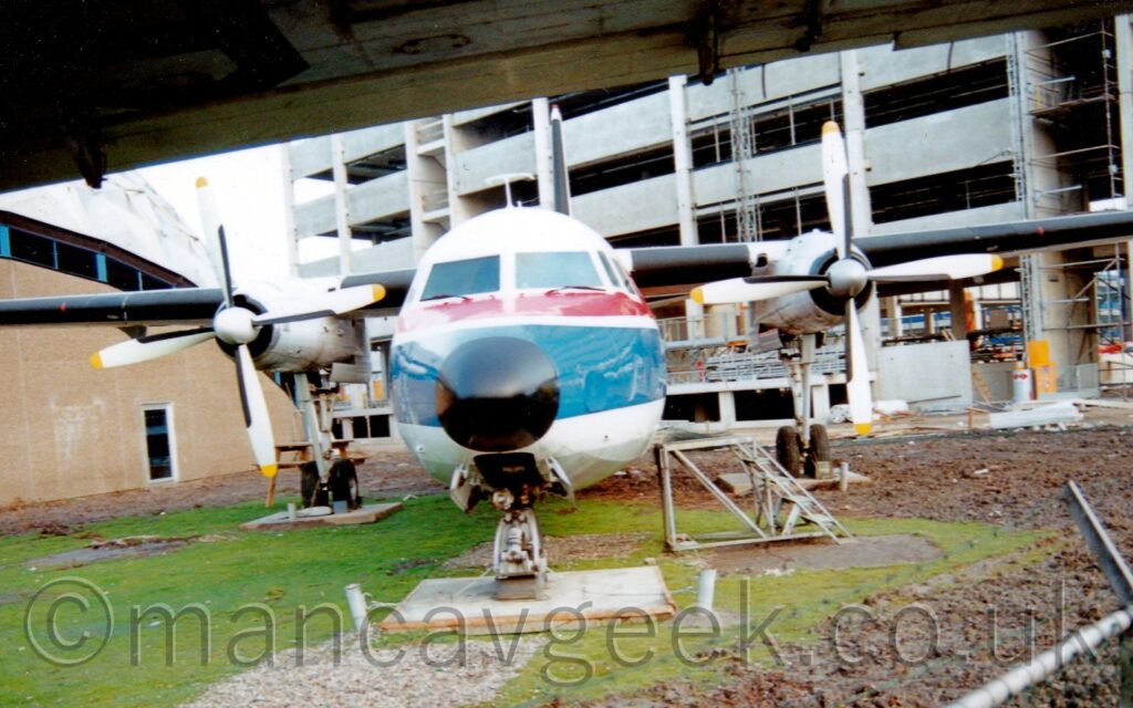 Head on view of a high-winged, twin propellor engined airliner facing straight down the camera lens, parked on concrete slabs in the middle of a grassed area.. The plane is mostly white, with a thin red stripe running along the body, just above a thicker blue stripe, both stripes just below the passenger cabin windows. A sandy brown building with a silvery domed roof is in the background on the left, with another, taller grey building is under construction on the right.