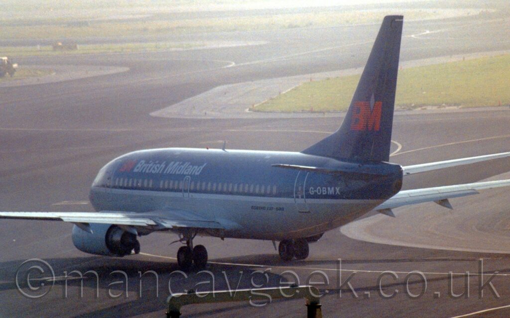 Side view of a twin engined jet airliner taxiing from right to left, following a yellow line along a black taxiway. The plane has a dark blue top section and a grey lower half, with a white stripe separating the 2 colours. There are white "British Miidland" titles on the upper forward fuselage, just aft of a red "BM" logo. The same logo is repeated in a larger form on the dark blue tail. The white registration "G-OBMX" is on the upper rear fuselage. Black taxiways wend their way through the background, skirting green islands of grass surrounded by grey concrete coastlines, slowly vanishing into haze as they move into the distance.
