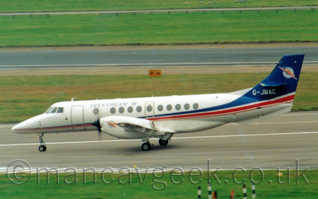 Side view of a twin propellor-engined airliner taxiing from right to left along a grey taxiway. The plane is largely white, with a thin red and blue stripe starting low down on the nose, getting wider and moving upwards as it moves rearwards, eventually the upper (blue) portion sweeping up to fill the tail. There are black "Jetstream 41" titles on the upper forward fuselage, and the white registration "G-JMAC" on the base of the tail. Green grass lines the side of the taxiway in the foreground, with more grass leading up to a black runway in the background, and more grass beyond that in the distance.
