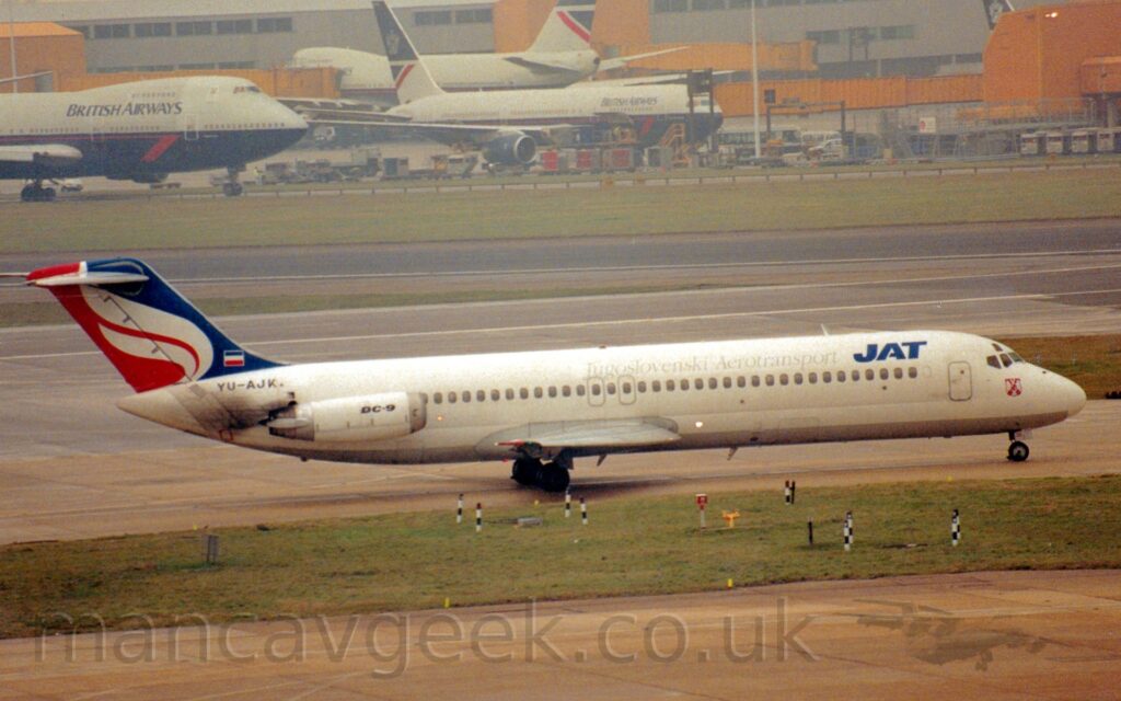 Side view of a twin engined jet airliner with the engines mounted on the sides of the rear fuselage, taxiing from left to right. The plane is almost entirely white, with bold, dark blue "JAT" titles on the upper forward fuselage, just ahead of thinner, grey, "Jugoslovenski AeroTransport" text. The black registration "YU-AJK" is on the upper rear fuselage, under a small Yugoslavian flag on the tail, while the white engine pod on the side of the rear fuselage has black "DC-9" text. The tail is mostly blue at the front and red st the rear, with something that is either a stylised white flame, or possibly a pair of wings, in the middle. A grey-brown taxiway and a wide strip of grass fill the foreground, while a grey building with yellow airbridges hanging off the side stretch across the background, with several grey and dark blue airliner parked in front.
