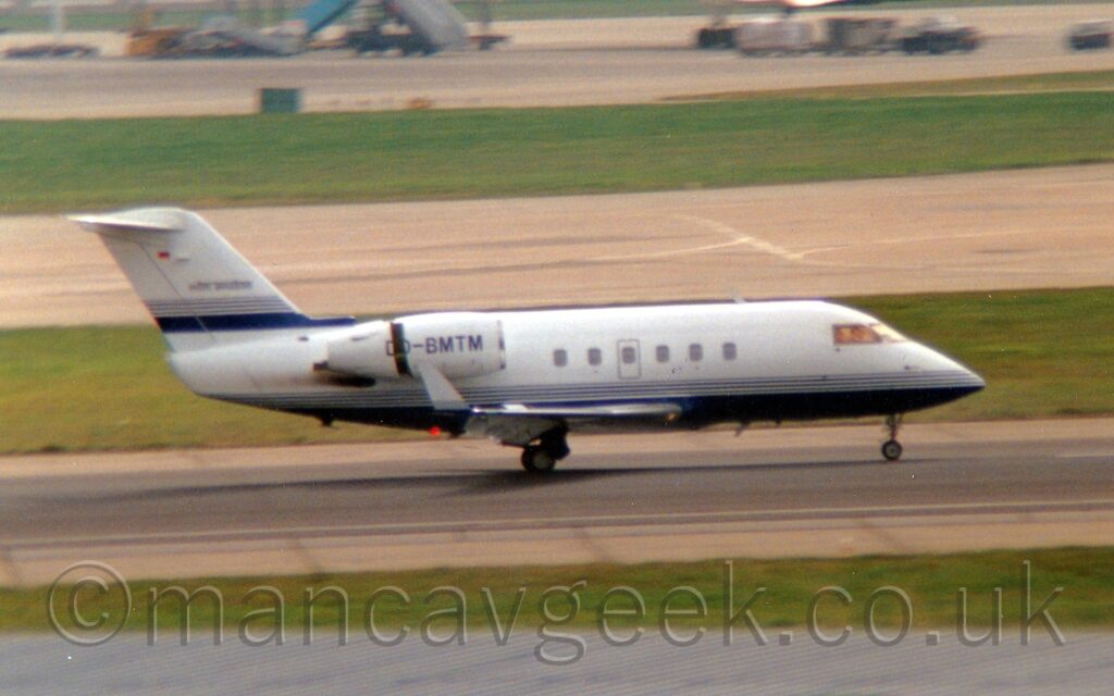 Side view of a twin engined BizJet moving from left to right at speed along a grey runway. Flaps are extended from the back of the wings, and the rear sections of the engine pods mounted on the sides of the upper rear fuselage are slid back, exposing the thrust reversers, suggesting that the plane is slowing down after landing. The plane is mostly white, with a dark blue belly and thin dark blue stripes along the lower fuselage. A similar thick dark blue stripe with thinner stripes on top is on the base of the white tail. The registration "D-BMTM" is on the side of the engine pod, the letter "D" being split on either side of the thrust reverser. A grey taxiway and a strip of grass line the side of the runway in the foreground, with more grass in the background giving way to a another taxiway, and more grass leading up to a large patch of apron in the distance.