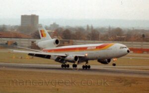 Front side view of a 3 engined jet airliner flying from left to right at a very low altitude, it's main gear about as close as you can be to a runway without actually touching it, with the nose-wheel just a bit higher up. The acres of flaps hanging from behind the wing, and the relatively shallow nose angle suggests the plane is in the process of landing. The plane is mostly white, with a brown, red, and yellow stripe coming down from the top of the forward fuselage, then along the body, covering most of the passenger cabin windows as it moves rearwards, overlaid with white "Iberia" titles on the upper forward fuselage. A stylised, red, lower-case letter "I" and yellow, uppercase letter "B" fill most of the tail above the centre-mounted engine, with a gold crown in the top part of the "I", and the black registration "EC-DEA" right at the top of the tail, repeated on the lower rear fuselage. Bright, red-tinged lights are shining in the wing root, and at the top of the nose-wheel leg. Yellowing grass fills the foreground, lining the side of the runway, with more grass in the background leading up to a high concrete wall, with trees and buildings beyond, under hazy grey sky.