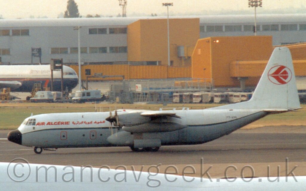 Side view of a high-winged, 4 propellor-engined cargo plane taxiing from right to left, crossing a black runway as it moves from one taxiway to another. The plane is white on top with a grey lower half, separated by a thin green stripe. There are red "Air Algerie" titles on the upper forward fuselage, in English at the front and in an Arabic script a bit further back. The black registration "7T-VHL" is on the upper rear fuselage. There is the red outline of a circle in the middle of the tail, containing a highly stylised red flying bird. The white fuselages of another pair of aeroplanes fills the foreground, with a wide strip of grass on the other side of the runway giving way to a grey building with large yellow airbridges filling the background, with the forward fuselage of a grey and blue parked on the left of the frame, all under flat grey sky.