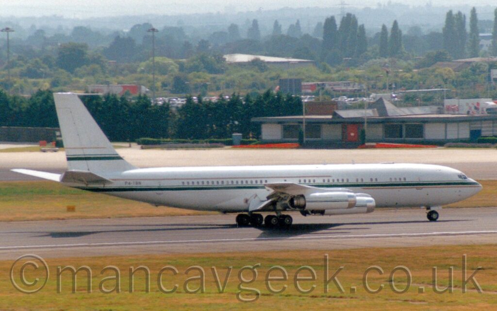 Side view of a 4 engined jet airliner moving at speed along a black runway, with flaps draped down behind the wings, suggesting it is slowing down after landing. The plane is mostly white with an green stripe running along the body under the passenger cabin windows, with a gold stripe under the green stripe in front of the wings, and above the windows aft of the wings. The green stripe with the gold stripe above is repeated on the lower part of the tail, and along the engine pods under the wings. Large grassed areas line both sides of the runway at the bottom of the frame, with a large area of concrete apron in the background leading up to a low grey building on the right, with trees in the distance stretching out to meet the grey skyline at the top of the frame.