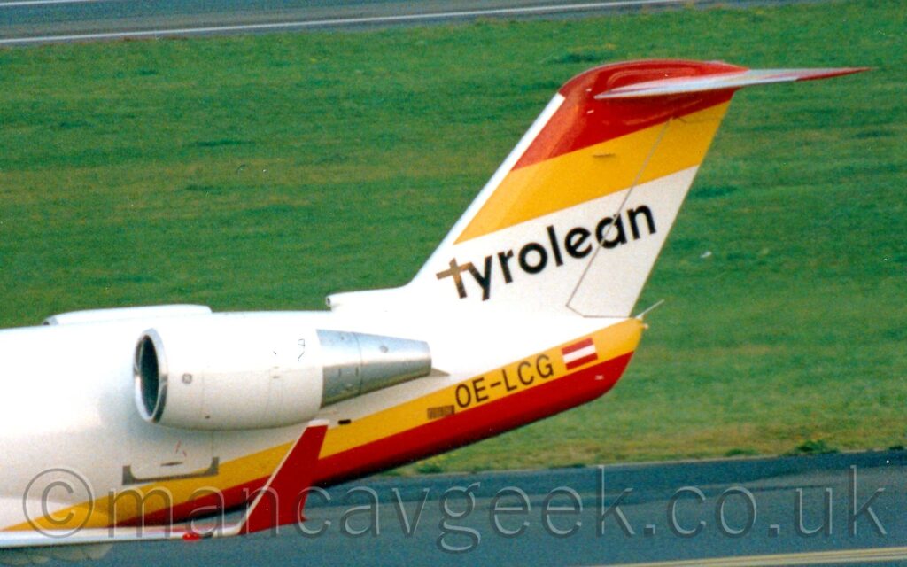 Close-up of the rear fuselage and tail of a twin engined jet airliner with the engines mounted on the sides of the rear fuselage, taxiing from right to left. The plane is mostly white, with a yellow, red, and brown striped belly, with those colours reversed at the top of the tail. There are diagonal black "Tyrolean" titles on the white portion of the tail, with the black registration "OE-LCG" next to a small, red and white Austrian flag on the yellow stripe on the tailcone. The engine pods on the sides of the rear fuselage are white, with chrome exhaust. The planes up-turned, red wingtip can be seen right at the bottom of the frame. Black tarmac taxiway and green grass fills the background, with the black runway visible in the top left corner.