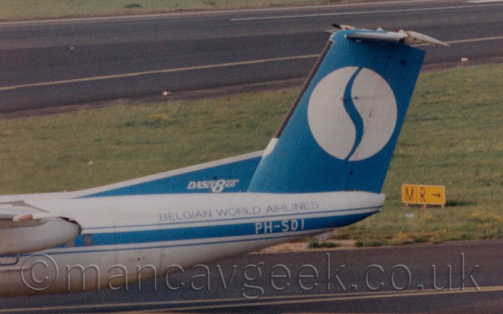 Close-up side view of the blue tail and white rear fuselage of a twin propellor-engined airliner taxiing from right to left. There is a blue stripe running along the body, covering the passenger cabin windows, and overlaid with the white registration "PH-SDI" under the tail, with blue text "Belgian World Airlines" just above. There is white text "Dash8" on a blue fillet at the front of the tail, with smaller text "Series 300" immediately after. The tail itself has a large with circle in the middle, with a stylised blue letter "S" that looks more like a slash. Black taxiway fills the foreground, with a large section of green grass and a black runway in the background.