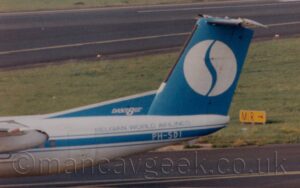 Close-up side view of the blue tail and white rear fuselage of a twin propellor-engined airliner taxiing from right to left. There is a blue stripe running along the body, covering the passenger cabin windows, and overlaid with the white registration "PH-SDI" under the tail, with blue text "Belgian World Airlines" just above. There is white text "Dash8" on a blue fillet at the front of the tail, with smaller text "Series 300" immediately after. The tail itself has a large with circle in the middle, with a stylised blue letter "S" that looks more like a slash. Black taxiway fills the foreground, with a large section of green grass and a black runway in the background.