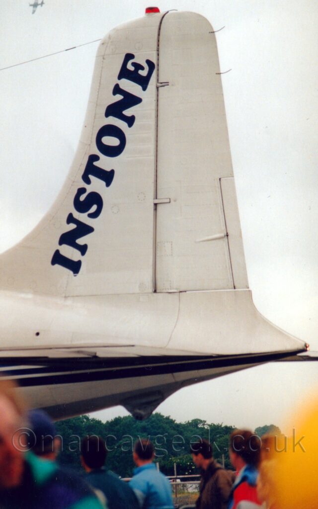 Close-up of the white tail of a vintage piston-engined airliner parked facing to the right. The plane is mostly white, with a grey belly, and a pair of thin black stripes running along the body. Diagonal black "Instone" titles run along the leading edge of the tail. Several people wearing padded coats a re standing in the foreground, most of them looking up at this plane. Trees are visible in the distance under the plane, with flat white sky filling the rest of the frame.